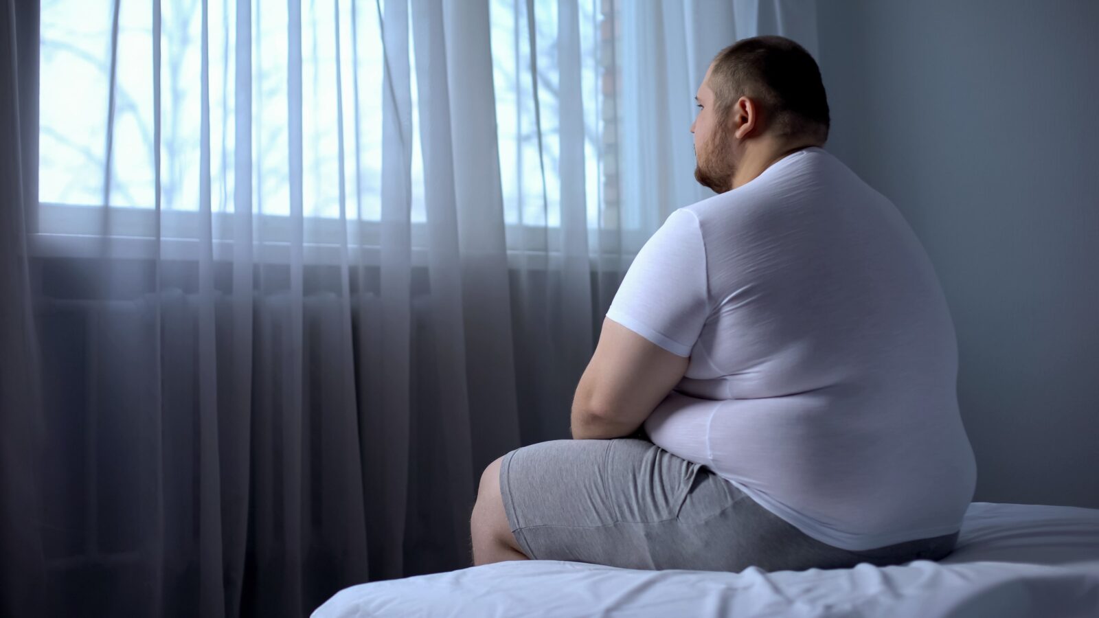 obese man sitting on bed and looking out the window